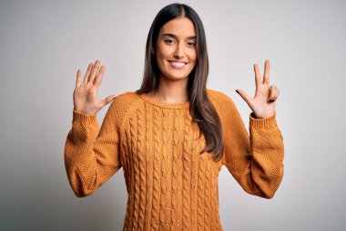 Genç, güzel esmer kadın izole edilmiş beyaz arka plan üzerine günlük süveter giyer ve 8 numaralı parmaklarıyla işaret ederken kendinden emin ve mutlu gülümserdi..