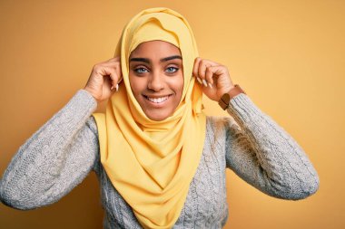 Genç ve güzel Afro-Amerikalı kız izole edilmiş sarı arka planda Müslüman başörtüsü takıyor parmaklarıyla kulaklarını çekerek gülüyor, komik bir jest. Seçme sorunu