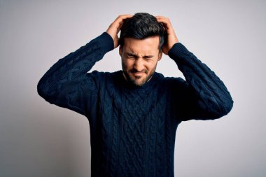 Sakallı, sıradan bir kazak giyen, beyaz arka planda dikilen, baş ağrısı çeken, çaresiz ve stresli genç bir adam. Çünkü acı ve migren. Ellerini başının üstüne koy..