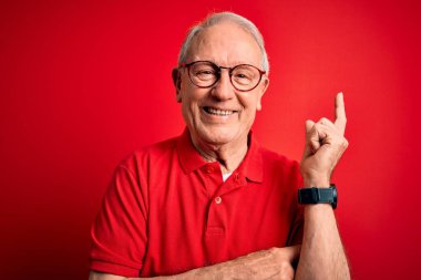 Gri saçlı, gözlüklü ve kırmızı arka planda rahat tişörtlü, yüzünde kocaman bir gülümseme olan, eli ve parmağı kameraya bakan son sınıf öğrencisi..