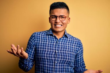 Sarı arka plan üzerine günlük gözlük takmış genç, yakışıklı latin adam güler yüzlü, güler yüzlü, dost canlısı, olumlu ve kendinden emin selamlar.