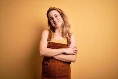 Sarı arka planda dikilip kollarını kavuşturup kameraya bakarken gülümseyen güzel sarışın kadın. Pozitif kişi.