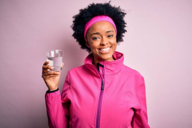 Genç Afrikalı Afro Amerikalı kıvırcık saçlı, bir bardak su içen, mutlu yüzlü, kendine güvenen, gülümseyen, dişlerini gösteren bir kadın.