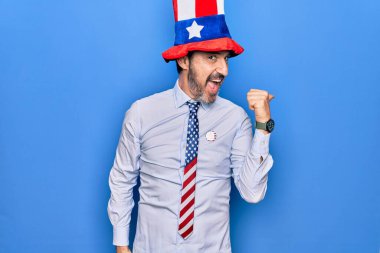 Orta yaşlı, yakışıklı, vatansever, şapkalı ve kravatlı bir adam mavi arka planda başparmağını yukarı kaldırıyor ve ağzı açık gülümsüyor.