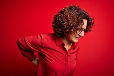 Orta yaşlı, kıvırcık saçlı, rahat gömlekli, kırmızı arka planda gözlüklü güzel bir kadın sırt ağrısı çekiyor, elleriyle dokunuyor, kas ağrısı çekiyor.