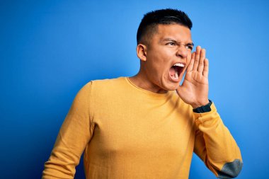 Sarı günlük süveter giyen genç, yakışıklı latin adam izole edilmiş mavi arka planda avazı çıktığı kadar bağırıyor ve el pençe divan bağırıyor. İletişim konsepti.