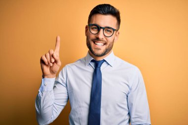 Sarı arka plan üzerinde duran kravat ve gözlük takan genç yakışıklı işadamı bir numaralı parmağını gösterip gülümserken kendinden emin ve mutlu görünüyor..