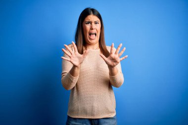 Mavi arka plan üzerine sıradan süveter giyen genç ve güzel bir kadın korku ifadesi karşısında korkmuş ve dehşete düşmüş. El hareketlerini durdur, şok içinde bağır. Panik konsepti.