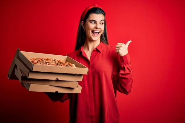 年轻美丽的黑发女子拿着装有意大利披萨的送货盒 背对着红色的背景 手指朝外 脸上挂着快乐的笑容 — 图库照片