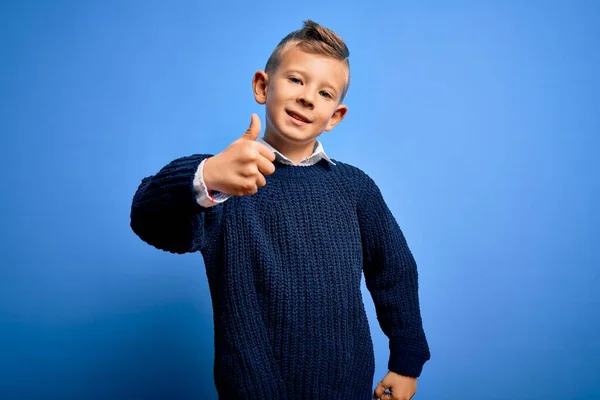 年轻的高加索小孩 蓝眼睛 穿着冬季毛衣 蓝色背景 手举着快乐的大拇指 在镜头前看着成功的表情 — 图库照片