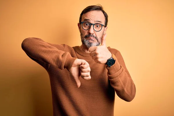 Middle age hoary man wearing brown sweater and glasses over isolated yellow background Doing thumbs up and down, disagreement and agreement expression. Crazy conflict