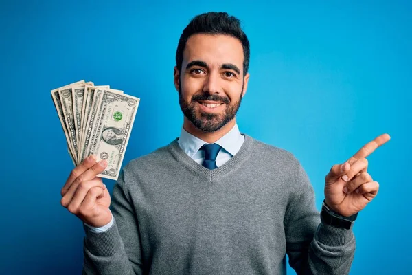 年轻英俊的男人留着胡子 手里拿着一堆钞票 背景是蓝色的 他非常高兴地把手指和手指头指向旁边 — 图库照片