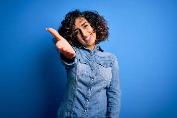 年轻美丽的卷曲阿拉伯女人穿着休闲斜纹棉布衬衫站在蓝色背景上 友好地握手致意和欢迎 成功的业务 — 图库照片