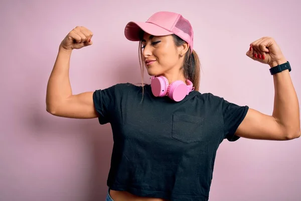 年轻美丽的健身女运动员戴着训练帽 戴着粉色背景的耳机 露出胳膊肌肉的笑容 感到自豪 健康概念 — 图库照片