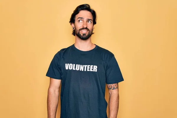 ボランティアのTシャツを着た若いハンサムなヒスパニック系のボランティアの男性は 横を見て笑顔と離れて考えて見つめて — ストック写真