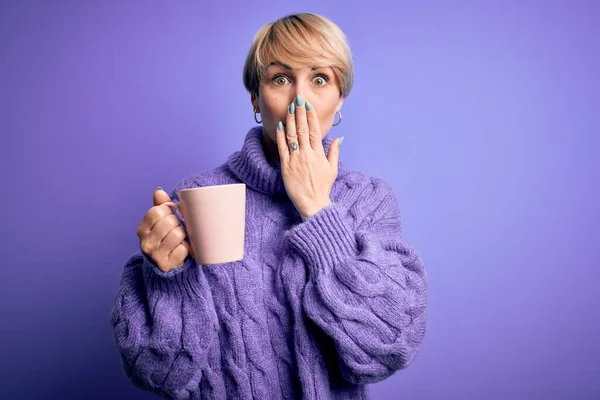 Young blonde woman with short hair wearing winter sweater drinking a cup of hot coffee cover mouth with hand shocked with shame for mistake, expression of fear, scared in silence, secret concept