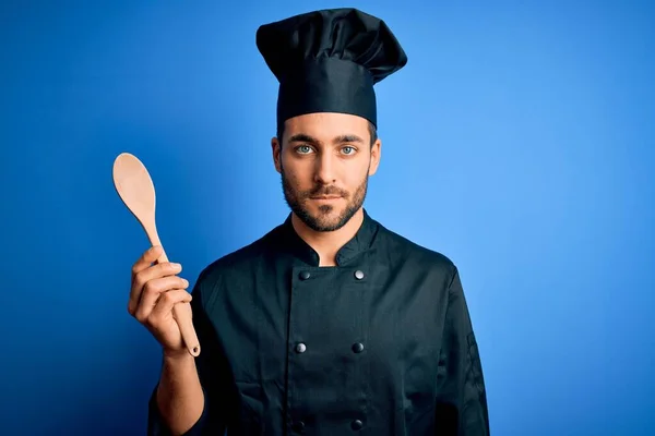 年轻的炊事员 留着胡子 身穿制服 蓝色背景上拿着木勺 脸上带着自信的表情 严肃地思考着 — 图库照片