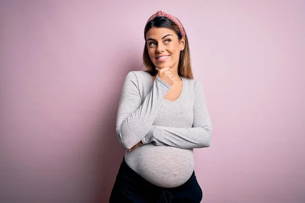若い美しいブルネットの女性は 孤立したピンクの背景の上に赤ちゃんを期待しています 思慮深い顔で微笑んだ 疑いの概念 — ストック写真