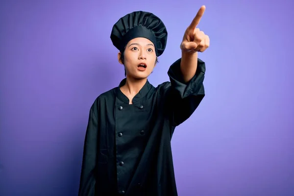 年轻美丽的中国厨师女士 身穿炊具制服 头戴紫色背景的帽子 手指尖尖尖 手指尖尖尖 满口惊讶的表情 前面有什么东西 — 图库照片