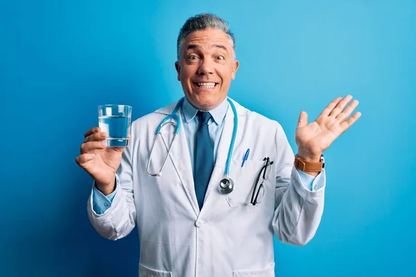 中年ハンサムな白髪の医師男は青の背景に水のガラスを飲んで非常に幸せと興奮し 勝者の式は大きな笑顔で叫んで勝利を祝うと手を上げ — ストック写真