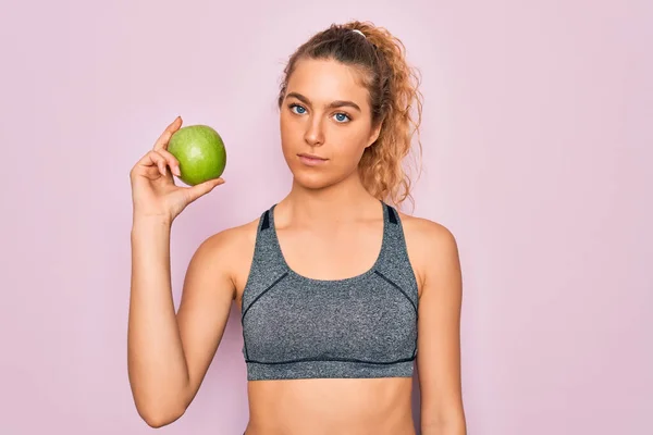 年轻美丽的金发女运动员 蓝眼睛 手持健康的绿色苹果果实 聪明的脸上带着自信的表情 严肃地思考着 — 图库照片