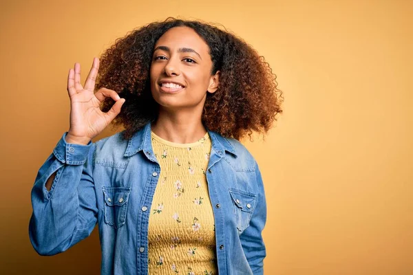 黄色の背景の上にカジュアルなデニムシャツを着てアフロの髪を持つ若いアフリカ系アメリカ人女性は 手と指でOkサインを行う肯定的な笑顔 成功した表現 — ストック写真