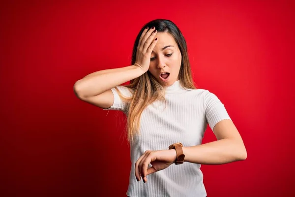Beautiful blonde woman with blue eyes wearing casual white t-shirt over red background Looking at the watch time worried, afraid of getting late