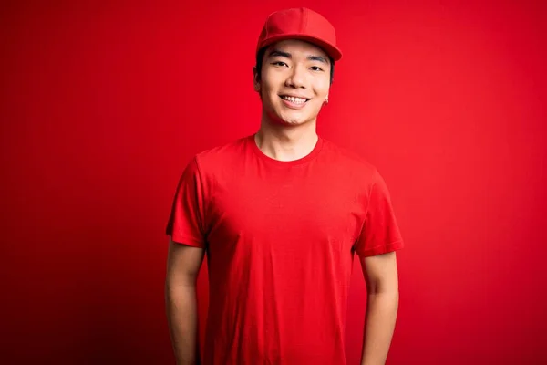 顔に幸せとクールな笑顔で孤立した赤い背景の上に立ってキャップを身に着けている若いハンサムな中国の配達人 運のいい人 — ストック写真