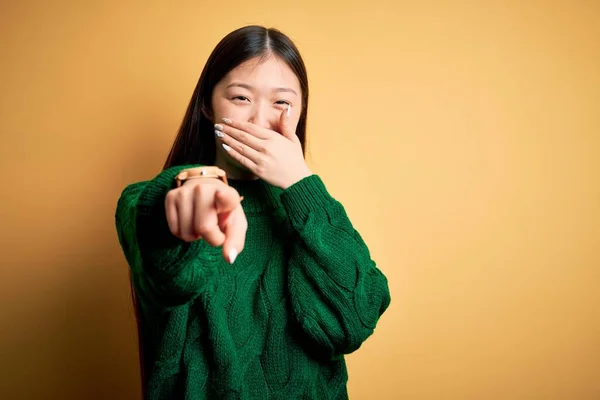 若いです美しいアジアの女性身に着けています緑の冬のセーター以上の黄色の隔離された背景あなたで笑って 口の上に手でカメラに指を指して 恥の表情 — ストック写真