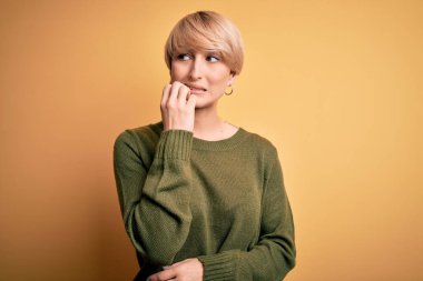 Modern kısa saçlı, sarı arka planda rahat süveter giyen genç sarışın kadın elleri ağız yiyen tırnaklarla gergin ve stresli görünüyor. Endişe sorunu.