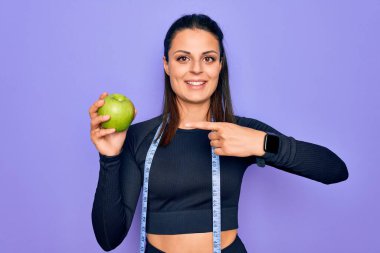 Güzel esmer sporcu kadın mezura kullanıyor mor arka planda elma meyvesi yiyor mutlu bir gülümsemeyle eli ve parmağı ile işaret ediyor