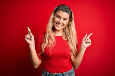 İzole edilmiş kırmızı arka planda dikilen ve parmaklarıyla farklı yönleri gösteren kendine güvenen gündelik tişört giyen genç, güzel sarışın kadın. Reklam için boşluk kopyala