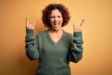 Orta yaşlı güzel kıvırcık saçlı kadın izole edilmiş sarı arka plan üzerine günlük süveter giyip çılgın bir ifadeyle elleri havada rock sembolü yaparak bağırıyor. Müzik yıldızı. Ağır konsept.
