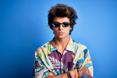 Gangster gözlüğü takan genç yakışıklı bir adam izole edilmiş mavi arka planda kuşkucu ve gergin duruyor. Yüzünde çapraz kollarla onaylamayan bir ifade var. Negatif kişi.