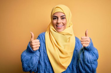 Genç, güzel, esmer, Müslüman kadın izole edilmiş sarı arka plan işareti üzerine Arap başörtüsü takıyor. Elleriyle olumlu jest yapıyor, gülümsüyor ve mutlu. Neşeli ifade ve kazanan jest.