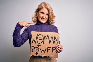 Orta yaşlı, güzel sarışın kadın kadın kadın hakları istiyor. Elinde pankart var ve kendini işaret ediyor.