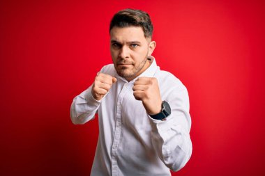 Mavi gözlü, zarif gömlekli genç bir iş adamı. Kırmızı izole edilmiş arka planda duruyor. Yumruk savunma hareketleriyle dövüşmeye hazır.