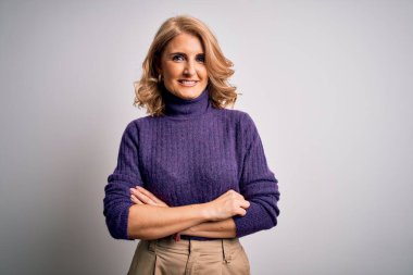 Beyaz arka planda mor balıkçı yaka kazak giyen, kollarını kavuşturup kameraya gülümseyen orta yaşlı güzel sarışın kadın. Pozitif kişi.