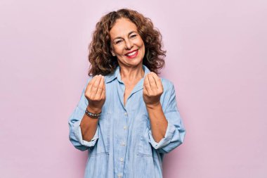 Orta yaşlı, güzel bir kadın pembe arka plan üzerinde dikilmiş para jestleri yapıyor, maaş ödemesi istiyor, milyonerlik işi yapıyor.