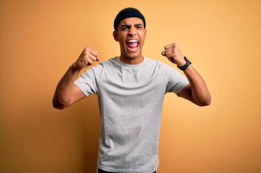 Yakışıklı Afro-Amerikan sporcu sarı arka plan üzerine spor giysisi giyiyor kızgın ve öfkeliyken kızgın yumruklar sallıyor ve öfkeyle bağırıyor. Öfke ve agresif kavram.