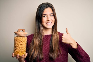 Genç ve güzel bir kız beyaz arka planda İtalyan makarnası şişesi tutuyor. Kocaman gülümsüyor.