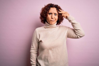 Orta yaşlı güzel kıvırcık saçlı kadın pembe arka plan üzerine balıkçı yaka kazak giyiyor. Alnındaki sorun yüzünden endişeli ve stresli. Kriz için endişeli.