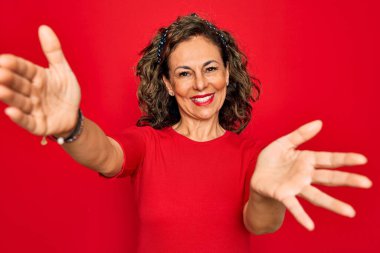 Orta yaşlı, esmer, sıradan tişörtlü bir kadın kırmızı arka planda dikiliyor ve kameraya sarılıp gülümsüyor. Mutluluğu kucaklayan neşeli bir ifade..