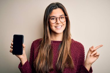 Gözlük takan genç ve güzel bir kız izole edilmiş beyaz arka planda akıllı telefon gösteriyor.