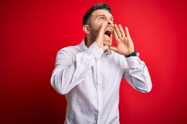 Mavi gözlü, zarif gömlekli genç bir iş adamı kırmızı izole arka planda dikiliyor ve elleri ağzında kızgın bir şekilde bağırıyor.