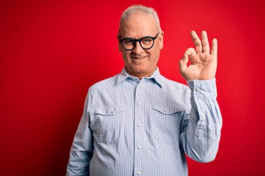 Orta yaşlı yakışıklı beyaz adam kırmızı arka planda günlük çizgili gömlek ve gözlük takıyor. Gülümsüyor ve el ve parmaklarıyla tamam işareti yapıyor. Başarılı ifade.