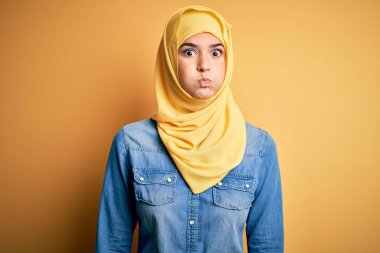 İslami tesettüre bürünmüş genç ve güzel bir kız izole edilmiş sarı arka planda durup komik yüzlü yanakları şişiriyor. Havayla şişmiş ağız, çılgın bir ifade..