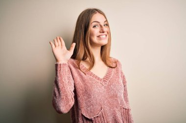 Genç, güzel, kızıl saçlı, pembe kazaklı, soyutlanmış beyaz arkaplanlı bir kadın. Mutlu, güler yüzlü, dostça bir jest yapıyor.