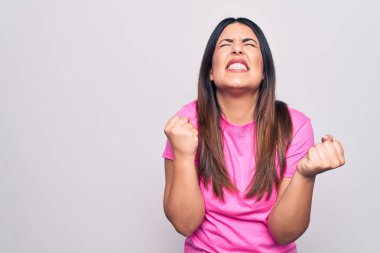 Genç, güzel, esmer, günlük pembe tişörtler giyen, beyaz arka plan üzerinde mutlu ve heyecanlı bir şekilde duran, kolları havada, gülümseyen ve başarı çığlıkları atan bir kadın. Kutlama konsepti.