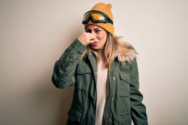 Genç esmer kayakçı kadın kar kıyafetleri ve kayak gözlüğü takıp beyaz arka planda kötü ve iğrenç bir koku alıyor. Nefesini burnunda tutuyor. Kötü kokuyor.